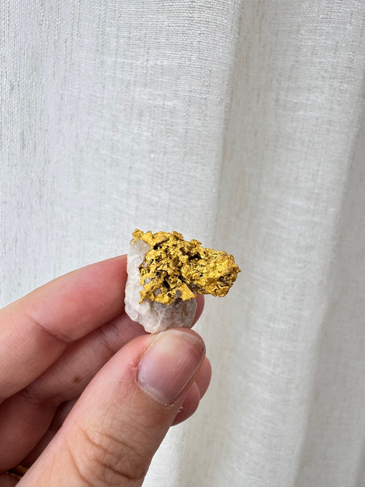 A hand holding a small piece of rough gold leaf and white stone against a textured off-white fabric background.