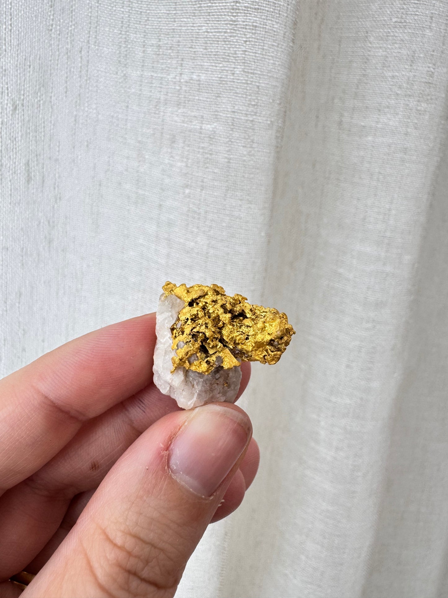 A hand holding a small piece of rough gold leaf and white stone against a textured off-white fabric background.