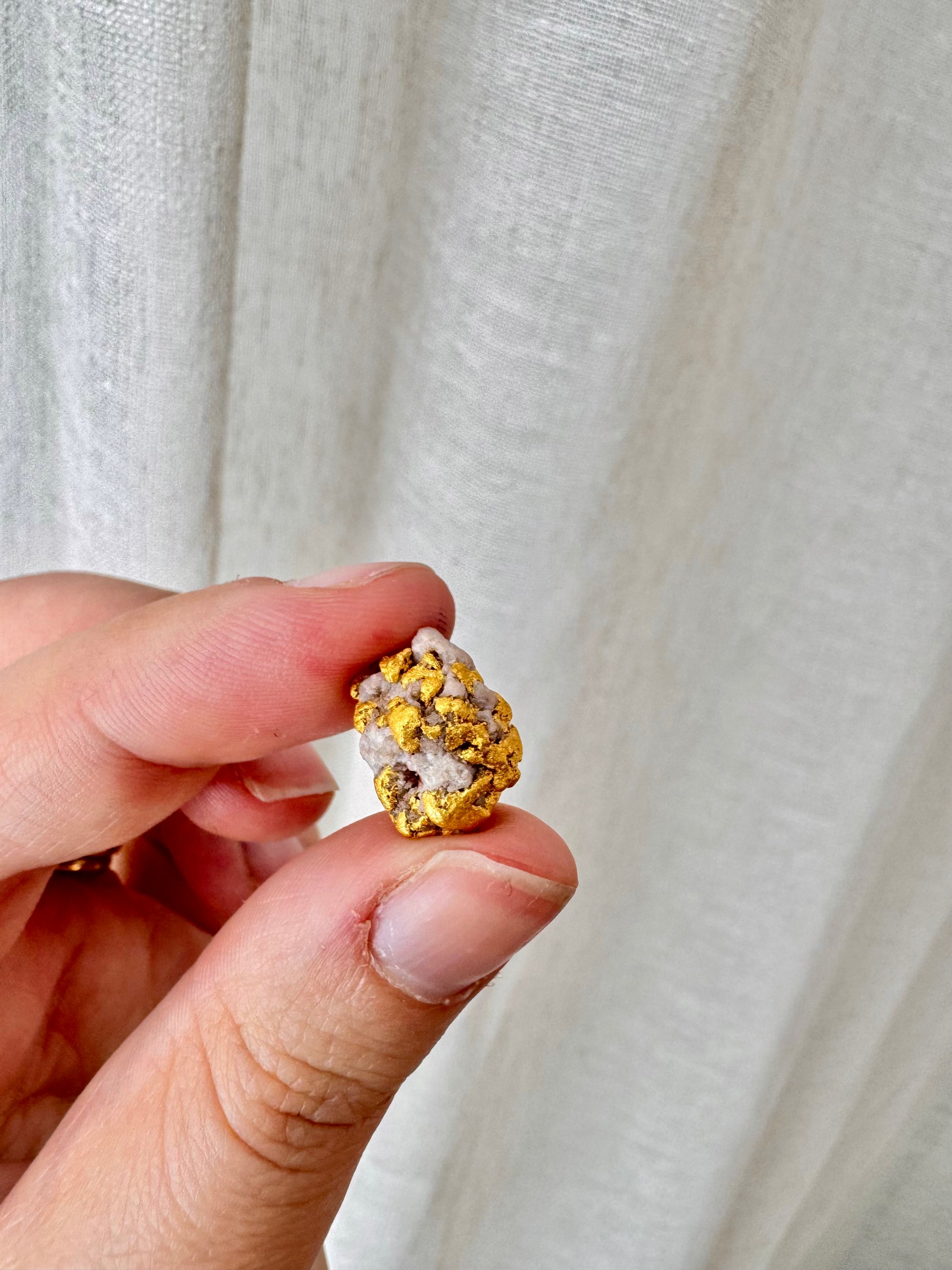 A hand holding a small gold and white textured jewelry piece against a soft, draped off-white fabric background.